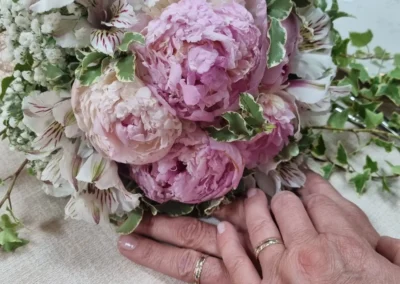 Dettaglio delle mani degli sposi con fedi d’oro accanto a un bouquet di peonie rosa e fiori bianchi, matrimonio in Abruzzo